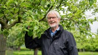 Der Baumwart Klaus Flaskamp, ein älterer Mann mit Brille und schwarzer Regenjacke, steht vor einem Obstbaum und hält einen Ast in seiner rechten Hand.