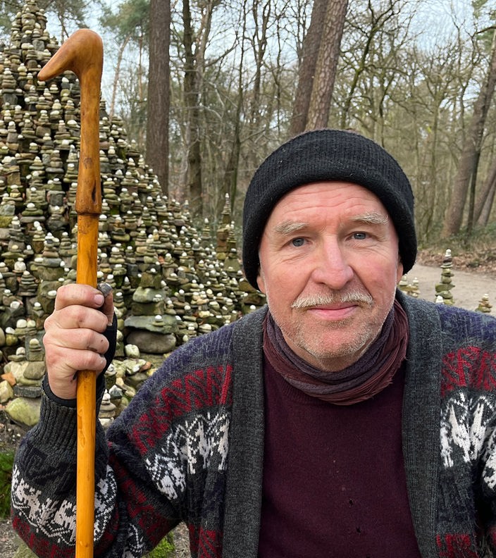 Künstler Markus Gern kniet im Steinmännchen-Wald in Kleve vor einem hohen Hügel aus Steinen