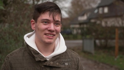 Teenager mit braunem Haar, weißem Hoodie und olivfarbener Jacke steht draußen vor einem Haus und gibt ein Interview