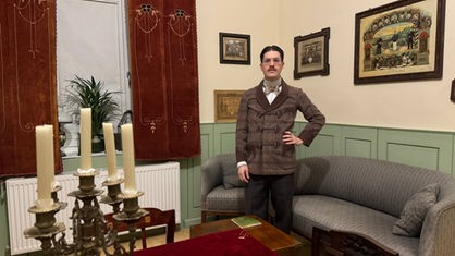 Ein Mann steht in einem Wohnzimmer, welches mit alten Möbeln eingerichtet ist.