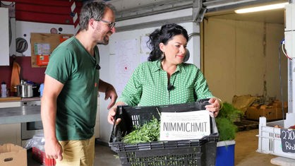 Zu sehen sind Marius Pötting und Chadia Hamadé, die eine Kiste hält, in welcher Salat und ein Schild mit dem Schriftzug "Nimm mich mit" liegt.
