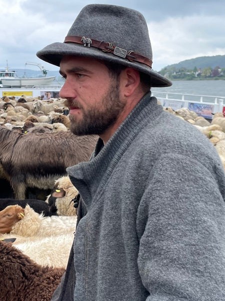 Mann in grauer Jacke mit grauem Filzhut auf dem Kopf steht inmitten von Schafen auf einer Fähre
