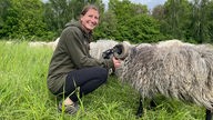 Claudia Lausberg sitzt in der Hocke schaut in die Kamera und streichelt gleichzeitig einen ihrer Schafböcke