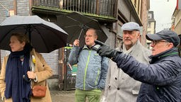 Man sieht vier Personen, zwei von ihnen tragen Regenschirme. Der Mann ganz recht zeigt auf etwas, in dessen Richtung die Blicke der Anderen folgen. 