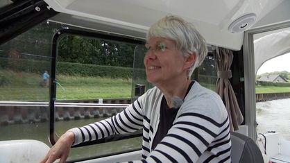 Eine Frau in weißem Cardigan mit schwarzen Streifen sitzt im Führerstand eines Motorboots und lächelt.