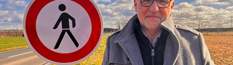 Auf dem Bild ist ein älterer Herr zu erkennen, der an einer Landstraße steht und direkt in die Kamera guckt. Neben ihm steht ein "Fußgänger verboten" Schild.