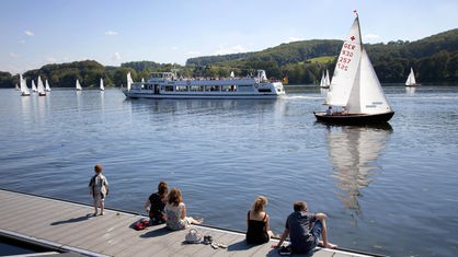 Junge Menschen sitzen am Baldeneysee auf einem Bootssteg in der Sonne. Vor ihnen Segelboote und ein Schiff mit Passagieren.