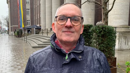 Ein Mann mit runder Brille, der in einer Regenjacke in die Kamera lächelt.