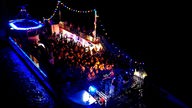 Drohnenaufnahme im Dunkeln von einem großen, bunt beleuchteten Schiff, auf dem eine Party stattfindet