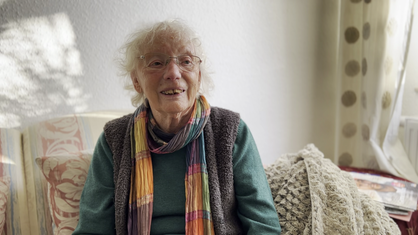 Eine Seniorin mit weißen Haaren und Brille sitzt auf einem hellen Sofa und lächelt. 