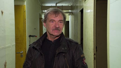 Mann in brauner Jacke steht in einem unterirdischen Bunker und spricht in Kamera 