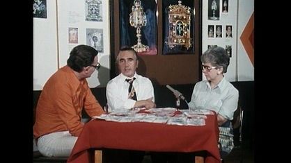 Zwei Männer und eine Frau sitzen an einem Tisch mit roter Tischdecke, auf dem ein Mikrofon steht