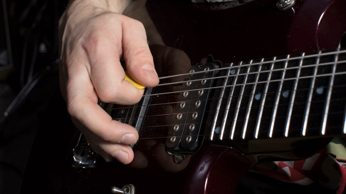 Nahaufnahme einer Person, die mit einem gelben Plektrum auf einer schwarzen E-Gitarre spielt.