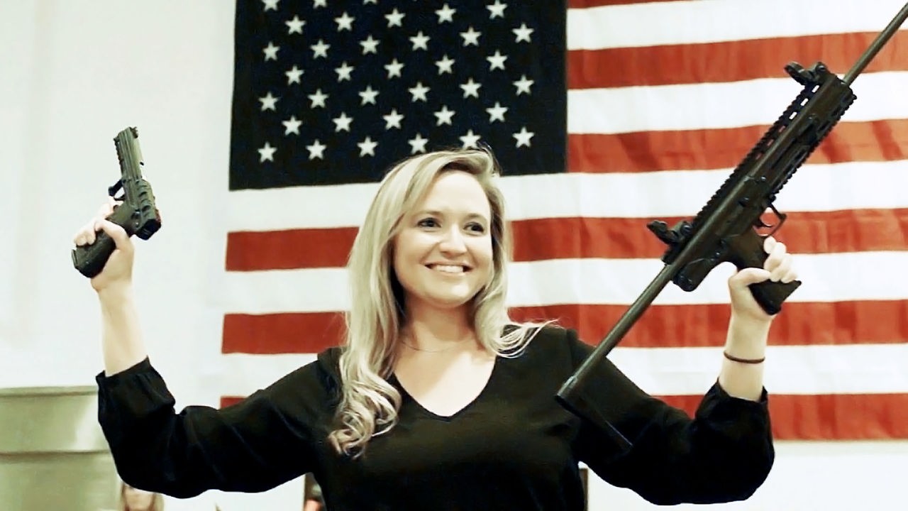 Eine Frau steht vor einer Amerika-Flagge und hält zwei Waffen in die Höhe.