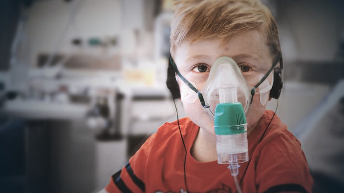 Ein kleiner Junge sitzt zentral in einem Raum eines Krankenhauses, trägt eine Sauerstoffmaske und schaut müde in die Kamera.