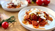 Das Gericht "Orecchiette mit Tomaten-Chorizo-Soße und Feta" in einem weißen Teller, drumherum liegen Tomaten und Kräuter.