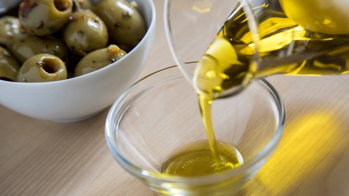 Olivenöl wird in eine Glasschale gefüllt während im Hintergrund eine Schale mit grünen Oliven steht.