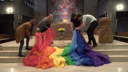 Vier Personen legen eine Regenbogenflagge in einer Kirche vor dem Altar ab.
