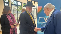 Goldschmidt ist mit schwarzem Hut und Karlspreis-Medaille um den Hals in der Aachener Synagoge