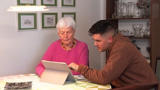 Ein junger Mann und eine Seniorein schauen auf ein Tablet.