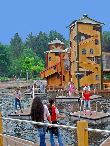 Ein großer Burg-ähnlicher Erlebnisspielplatz aus Holz ist umgeben von einem kleinen Wasserpark. Viele Besucher tummeln sich auf den Türmen des Spielplatzes und auf kleinen Floßen und Stegen.