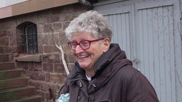 Lachende Frau mit kurzen grauen Haaren und Brille vor einem Haus