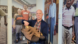 Georg Wessels steht vor einem Foto und hält Schuhe Größe 70 in der Hand