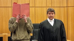 Eine Mann verdeckt sein Gesicht mit einem Ordner, neben ihm ein Anwalt.