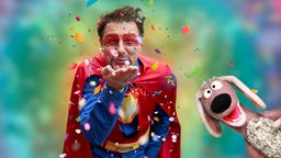 André Gatzke im Superman-Kostüm pustet Konfetti in die Kamera, der Dackl guckt rechts ins Bild