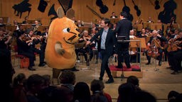 Johannes Büchs und die Maus stehen auf der Bühne, im Hintergrund das WDR Sinfonieorchester