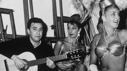 João Gilberto sitzt mit verschränkten Beinen auf einem Hocker und hält seine Gitarre. Neben ihm sitzen drei leicht bekleidete Tänzerinnen mit Perlenketten und Federkopfschmuck.