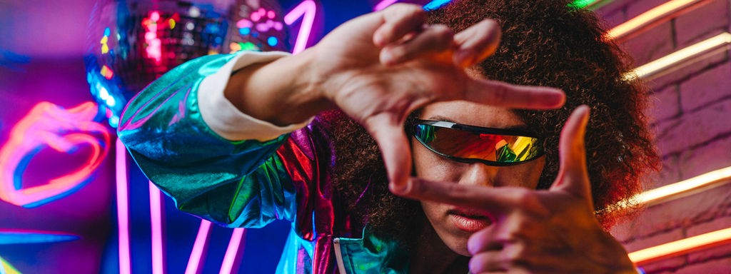 Frau mit futuristischer Brille formt ihre Finger zu einem Rahmen, hinter ihr Neonlicht.