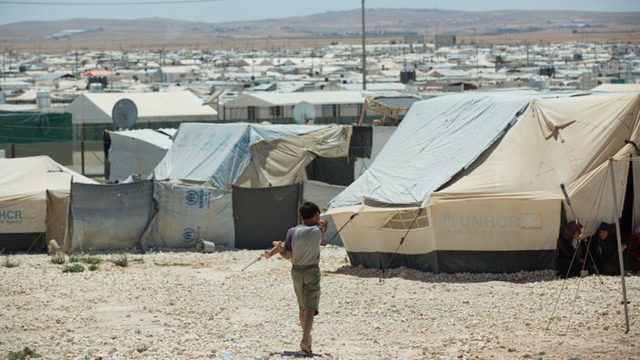 Ein Junge trägt ein kleines Kind auf dem Arm, dahinter ein Flüchtlingslager aus Zelten