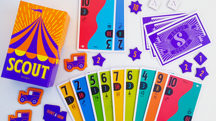 Die Spieleschachtel von "Scout" auf einem Tisch, daneben das Spielmaterial aus bunten Karten mit Werten von 1 bis 10 und Pappmarkern mit Zahlen darauf