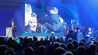 Blick aus dem Publikum auf die Bühne, Konfetti in der Lufr. Die Gewinner umarmen sich am 18.04.2024 bei der Preisverleihung. Das wird auf einer Leinwand noch mal groß übertragen.