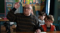 Michel Blanc in einer Szene des Films "Es sind die kleinen Dinge": Er sitzt zwischen Schülern in einer Klasse und zeigt auf.