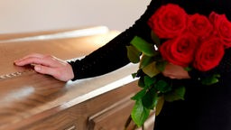 Eine Hand hält rote Rosen vor einem Holzsarg.