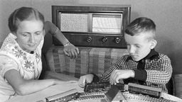 Mutter und Sohn hören im Wohnzimmer Radio, während das Kind mit einer Bahn spielt.