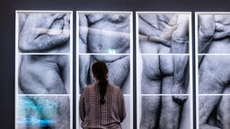Eine Fraut steht vor einem schwarz-weißen Fotos, die vier nackte Körper alter Menschen in sehr großem Format abbildet.