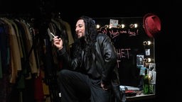 Bühnenszene aus "Ein von Schatten begrenzter Raum" am Schauspiel Köln, ein Mann mit langen schwarzen Haaren sitzt in einem augescheinlichen Dressing Room und hält einen Stift in der Hand.
