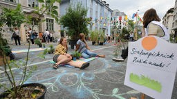 Symbolbild: Menschen sitzen neben Grünpflanzen auf einen bemalten, autofreien Straße, daneben ein Schild mit der Aufschrift "Straßenpicknick - Hock di nieder!"