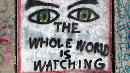 Ein Graffiti auf der Wand: zwei Augen mit der Unterschrift "the whole world is watching".