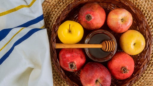 Äpfel und Granatäpfel in einem Korb, in ihrer Mitte glänzt ein Glas Honig