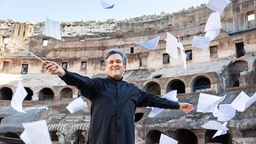 Der Dirigent Antonio Pappano im Collosseum in Rom, umgeben von Notenblättern.