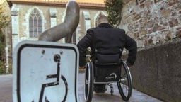 Symbolbild zu Barrierefreiheit bei Denkmälern: Ein Mann fährt in seinem Rollstuhl eine Rampe hoch, im Hintergrund eine alte Kirche.