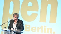 Deniz Yücel vor an einem Mikrofon, im Hintergrund der der Schriftzug des "pen Berlin".