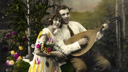Koloriertes Bildmotiv aus den 1920 Jahren: Mann und Frau im Grünen mit einer Laute und bunten Blumen.