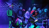 Szenenfoto von "Splash" am Marabu Theater Bonn: Vier Menschen mit Instrumenten und neonfarbenen Accessoires wie Taucherflossen oder Taucherbrillen bewegen sich auf einer blau beleuchteten Bühne.