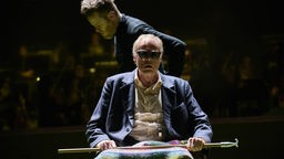Szenefoto von "Fin de Partie" am Theater Dortmund: Ein Mann mit Sonnenbrille und Hemd und knittrigem Anzug auf einem Stuhl unter einer Häkeldecke.