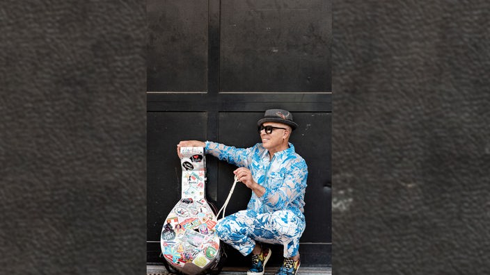 Der Musiker Dhafe Youssef sitzt in einem blau-weißen Anzug mit seiner Oud neben ihm in der Hocke.